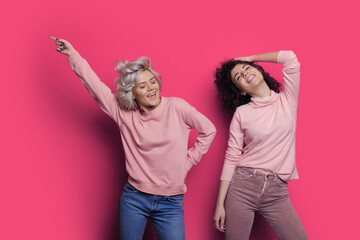Energetic two women dancing waving hands with smile on face, posing over pink background.