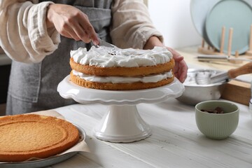 Woman smearing sponge cake with cream at white wooden table in kitchen, closeup