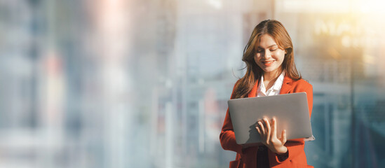 Pretty Asia business woman in red formal suit using laptop with blurry background wide picture layout.