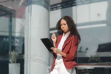 Fototapeta na wymiar Beautiful african american business woman in red formal suit leader entrepreneur, professional manager holding digital tablet, going to work, walking urban outdoor, standing on city street