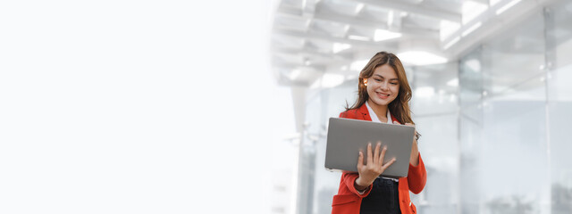 Using laptop, video call, Smiling young Asia business woman leader entrepreneur in red color suit working outdoor urban street walk.