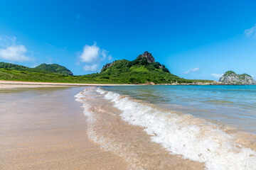 Onda correndo na areia e um morro verde ao fundo.