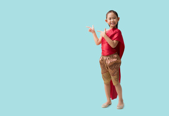 Full body Asian little girl in Thai national costumes with pointing finger blank workspace, isolated on pastel plain light blue background