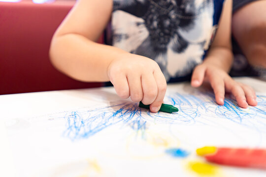 A child plays, studies, and learns how to color and draw with crayons on paper with the help of their parents. Kid's parents provide guidance and support as child develops their artistic skills.