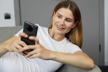 Pregnancy tracker app. Happy pregnant woman using smartphone at home. Smiling expectant mother reading information about baby development, laying on sofa with cellphone, relaxing  