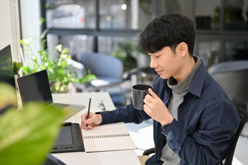 Attractive Asian male office worker concentrating on his task, making list on his notepad