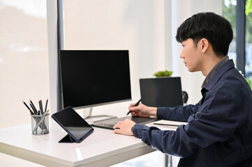 Professional Asian male website developer focusing on his project, using graphic tablet at his desk.