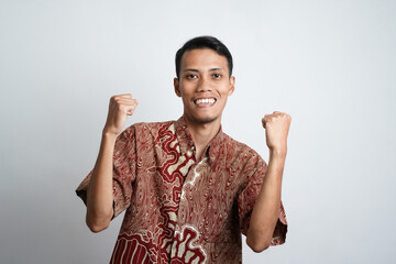 Excited Asian man wearing batik shirt showing strong gesture by lifting his arms and muscles...