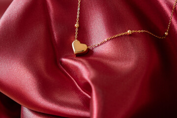 A gold necklace with a gold heart-shaped pendant resting on a pinkish-red fabric is the perfect...