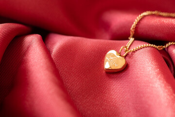 A gold necklace with a gold heart-shaped pendant resting on a pinkish-red fabric is the perfect...