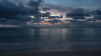 Calm evening seascape in shades of blue. Picturesque clouds in the sky, highlighted in pink. Reflection on the ocean surface. Foam of waves on wet sand. Long exposure. Seychelles. Mahe. Beau Vallon