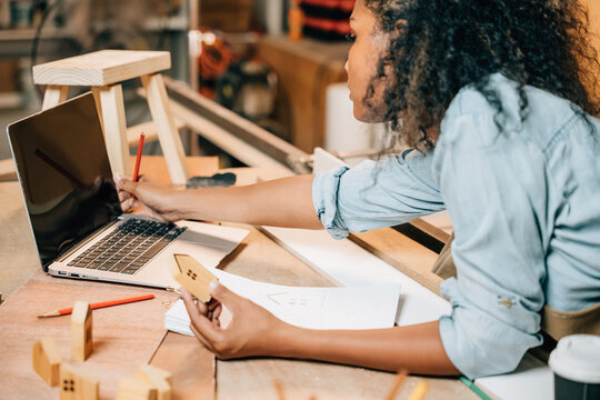 Carpenter America Black Woman Curly Hair Sketch Making Notes In Work Paper While Standing At Wooden Table With Laptop Computer, Young Female Working Learning Online At Woodshop, Happy Carpenters Day