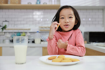 Kid asian black hair girl sitting on table and drinking milk while eating vanilla cookies for breakfast with enjoying time. Tasty food and delicious food with happy meal lifestyle kid concept