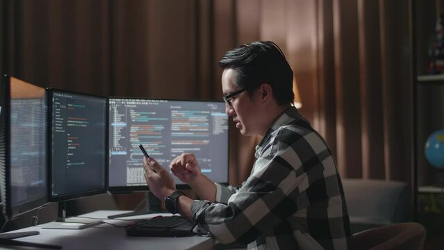 Asian Male Programmer Looking At Smartphone Then Celebrating The Success While Creating Software Engineer Developing App, Program, Video Game On Desktop Computer At Home
