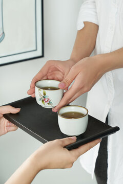 Shot of female hands holding a white porcelain color printed cup in chinese style. Male hands holding a wooden tray with the white cup are located on the background of a white wall with a painting.