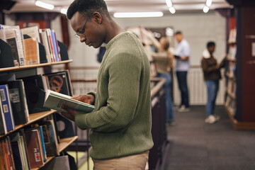 Black man student, reading and library with book, research and education at college to ready for exam. African gen z man, books and shelf at university for study, learning and motivation in Boston - obrazy, fototapety, plakaty