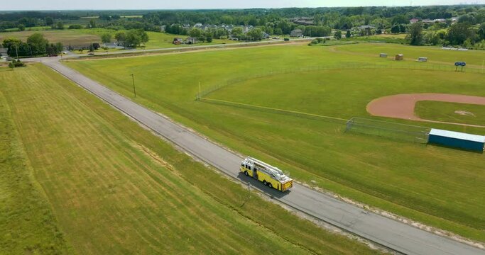 Yellow ladder firetruck backing up on street by fields – aerial view drone orbit