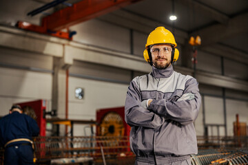 A confident metallurgy worker in protective work wear is standing in facility and smiling at the...