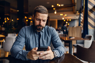 A serious man is sitting in a coffee shop with phone in his hands and reading e-mails.