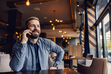 A smiling entrepreneur is sitting in coffee shop and talking on the phone with business partner.