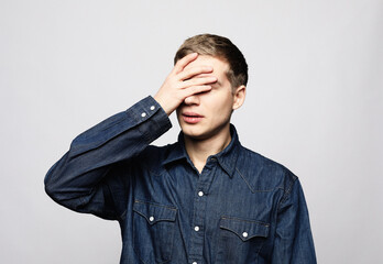 A young handsome man covers his eyes with his palm over grey background.