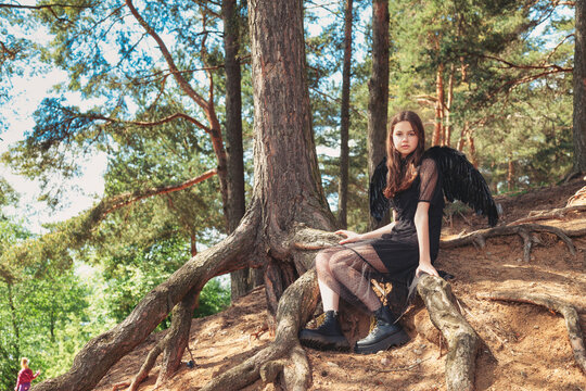 Teenager girl with long hair in black wings sitting in fairy tale woodland, looking at camera. Young lady Princess angel at mysterious forest. Vintage retro image, fantasy concept. Copy text space