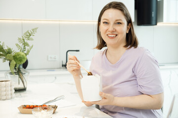Young plump woman sits in the kitchen and eats noodles from a paper white box. Food delivery. Unhealthy food.