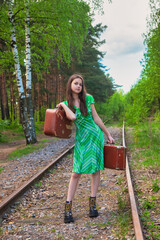 Teen girl in stylish retro image with vintage suitcases standing on railway in summer woodland, looking at camera. Fashionable young lady in green dress on forest. Travel vacation concept. Copy space