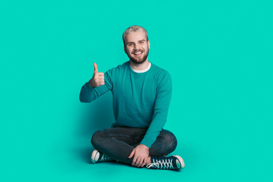 Full size photo of young man sitting on floor show thumbs-up approve isolated over blue color background. Positive person.