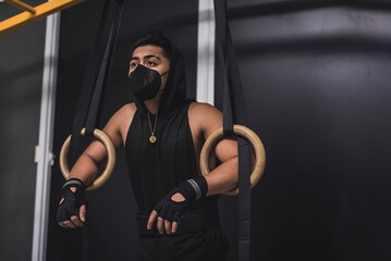 Fototapeta na wymiar A masked asian man geting in the zone leaning on wooden still rings. A gymnast preparing to train at the gym.