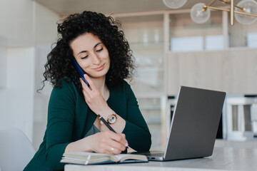 Successful young curly woman in green cardigan sitting at desk with laptop and notebook making call by phone looking at camera. Calm Spanish entrepreneur remote working at home. Business and finance.