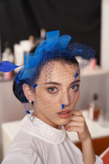 Cropped close-up shot of a young woman with a dark blue fascinator with a bow and a veil decorated with feathers. A girl in a white blouse and a fascinator is posing in the studio. Front view.