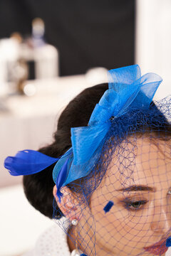 Cropped close-up shot of a young woman with a dark blue fascinator with a bow and a veil decorated with feathers. A girl in a white blouse and a fascinator is posing in the studio. Side view.