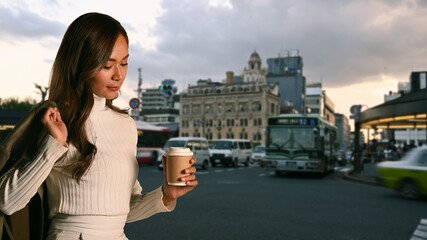 Trendy woman with paper up of coffee standing against background of busy city street and evening sky