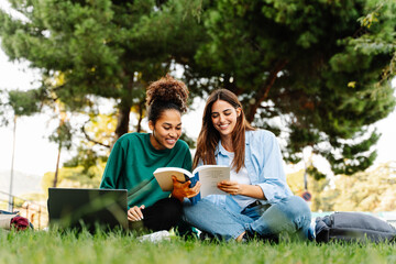 University student girl friends working and learning together using laptop sitting on campus grass