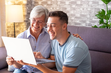 Young cheerful boy teaching his grandmother how to surf the internet, buy products online while...