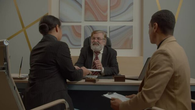Happy diverse multiracial business partners signing mutually beneficial contract and handshaking, expressing positive vibes, confidence in successful partnership during meeting at office.
