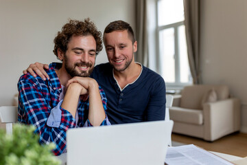Fototapeta premium Lovely gay couple laughing together while sitting in their living room at home. Two romantic young male lovers having fun surfing the internet indoors. Young gay couple living together.