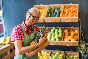 Mature woman works in fruits and vegetables shop. She is holding pineapple.