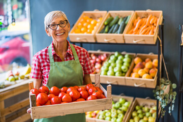 Mature woman works in fruits and vegetables shop. She is holding basket with tomatoes.