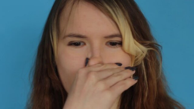 Close-up of a young Caucasian girl on a blue isolated background, smells an unbearable smell, holds her breath, pressing her fingers to her nose, an unpleasant smell.