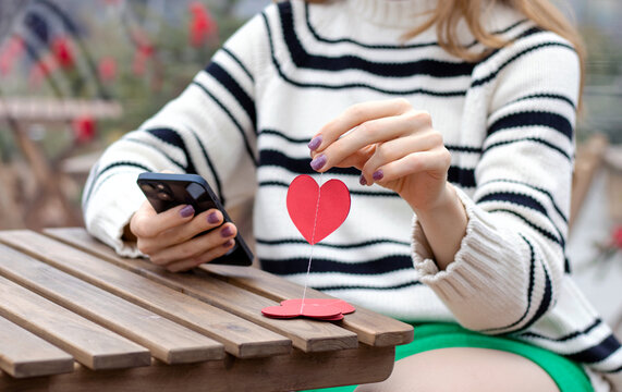 woman sitting in on table with paper hearts garland in one hand using smartphone with another,girl hugging red balloon heart shape.celebrate love valentine day coffee shop inside transparent balloon