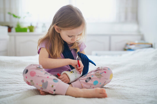 Adorable preschooler girl with a broken arm at home on the bed draws with felt-tip pens on an orthopedic cast