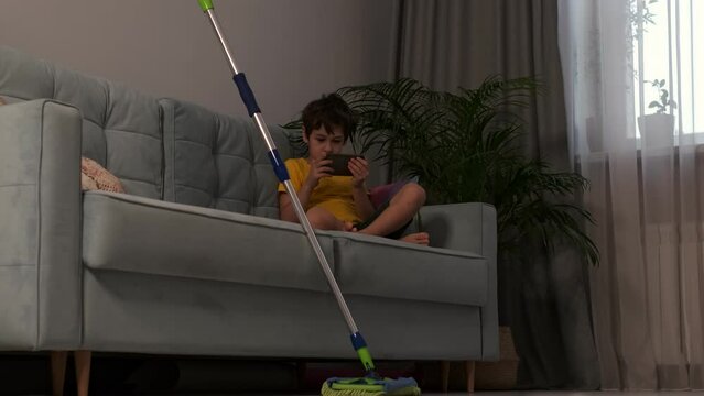 child sitting on the sofa and plays on the phone, an abandoned mop lies next to him. how to get a child interested in home cleaning concept