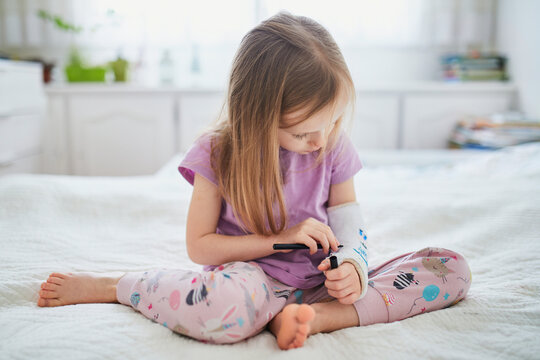 Adorable preschooler girl with a broken arm at home on the bed draws with felt-tip pens on an orthopedic cast