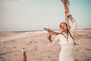 Model in boho style in a white long dress and silver jewelry on the beach. Her hair is braided, and there are many bracelets on her arms.