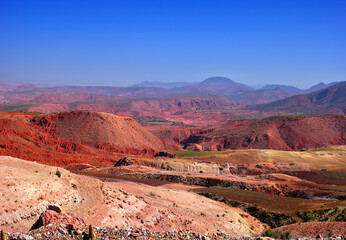 Beautiful desert view, barren multicolored hills in red, pink, purple and yellow colors, few green fields, distant mountains and blue sky in Southern Morocco, North West Africa.