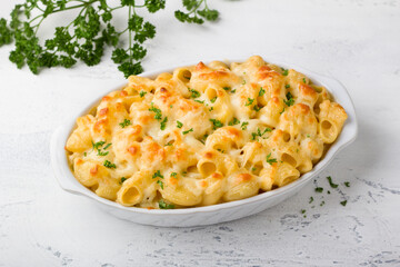 Mac and cheese, baked American pasta in cheese sauce decorated with parsley in a baking dish on a light gray background. Delicious homemade food