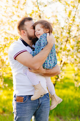 Dad hugs and kisses his little daughter on a walk in a blooming spring garden.