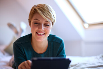 Young Smiling Woman Lying On Bed Wearing Pyjamas At Home Looking At Digital Tablet